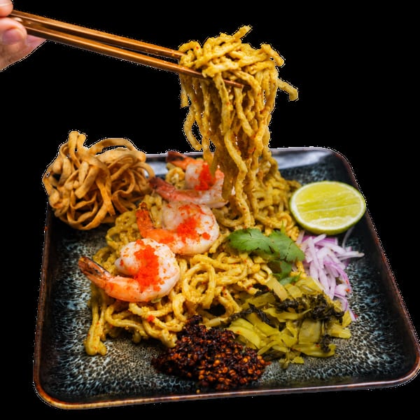 Chopsticks lifting golden noodles from a plate of shrimp khao soi with lime, onions, and chili.