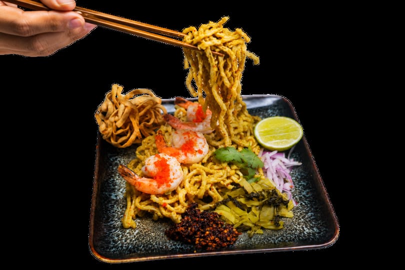 Chopsticks lifting golden noodles from a plate of shrimp khao soi with lime, onions, and chili.